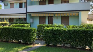 A two-story building with a light green facade, yellow-striped awnings, and a small garden area with neatly trimmed bushes. - Apartment With Garden and Pool Access (San Michele al Tagliamento)