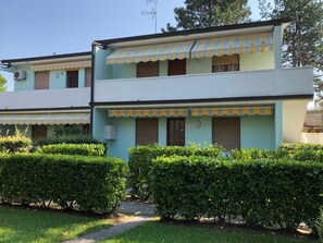 A two-story building with a light green facade, yellow-striped awnings, and a small garden area with neatly trimmed bushes. - Apartment With Garden and Pool Access (San Michele al Tagliamento)