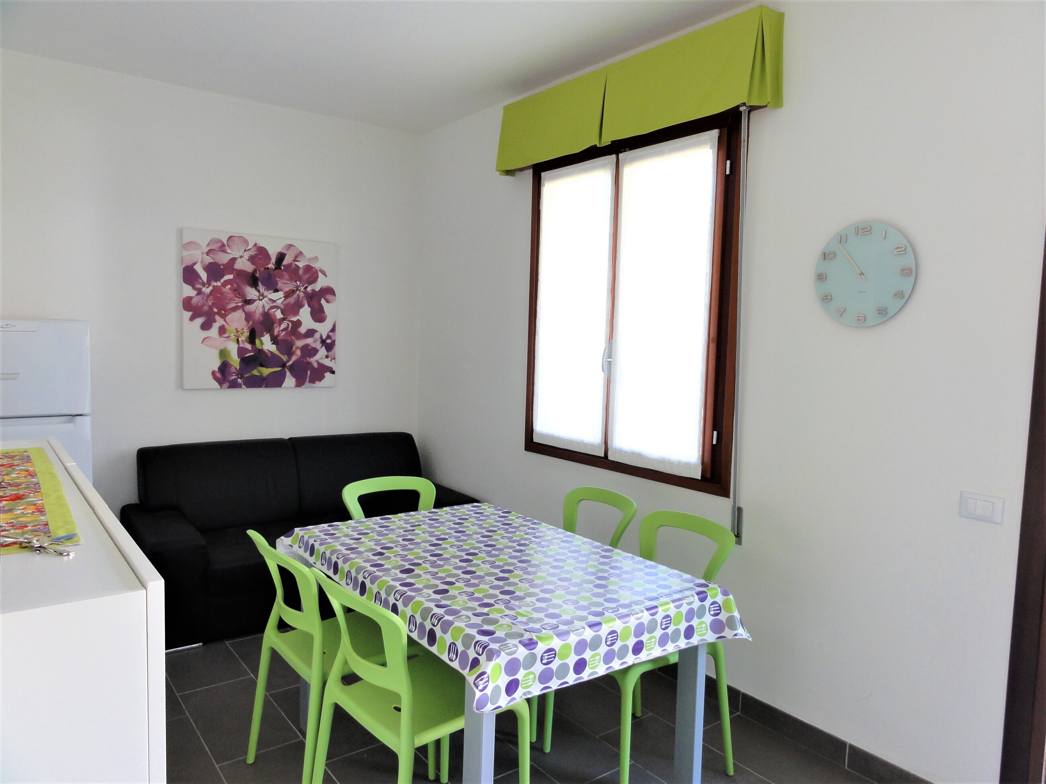 A small, bright room with a table, chairs, a sofa, a window with curtains, a clock, and a floral artwork on the wall.