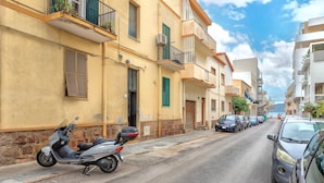 A street with parked cars and a scooter, buildings with balconies, and a clear sky. - Piera's Apartment In Alghero – Steps from the Seafront, Ideal for Couples and Families (Alghero)