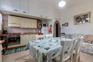 Une cuisine compacte avec des meubles blancs, un four à micro-ondes et une cuisinière. On y trouve une table à manger avec des chaises et une nappe à motifs. Un canapé à imprimé floral se trouve dans l’espace salon.