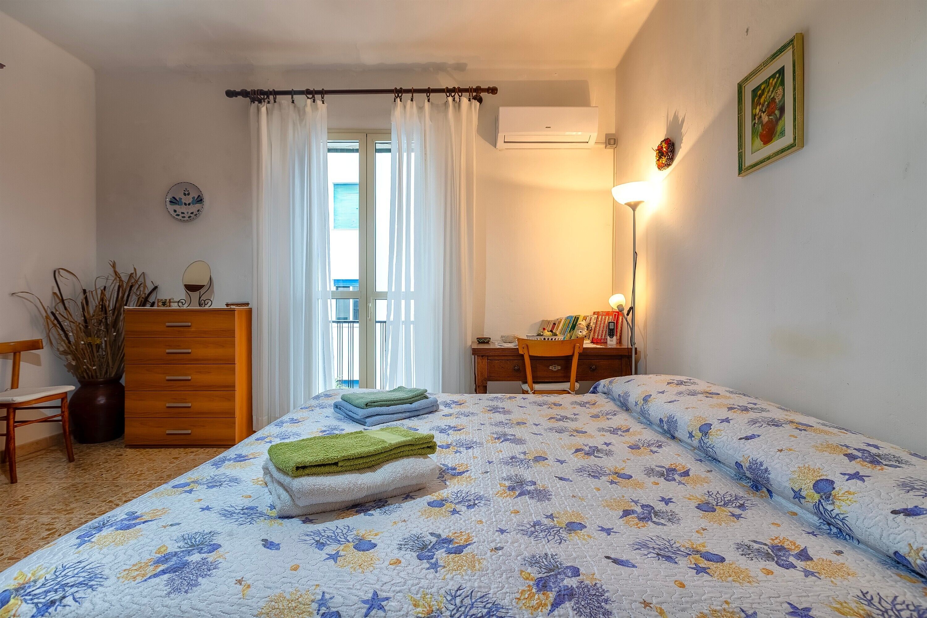 A bedroom with a bed, a dresser, a desk, a chair, a potted plant, and a window with curtains.