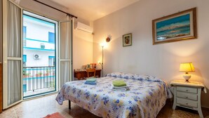 A bedroom with a bed, a nightstand, a lamp, a painting, and a view through a sliding door. - Piera's Apartment In Alghero – Steps from the Seafront, Ideal for Couples and Families (Alghero)