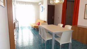 Un coin repas avec une table et des chaises blanches, un sol carrelé de bleu, un lit recouvert d’une couverture jaune et un rideau rouge.