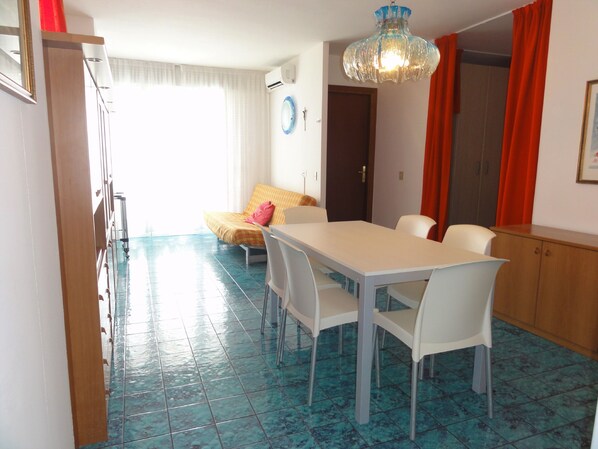 A dining area with a white table and chairs, a blue tiled floor, a bed with a yellow bedspread, and a red curtain.