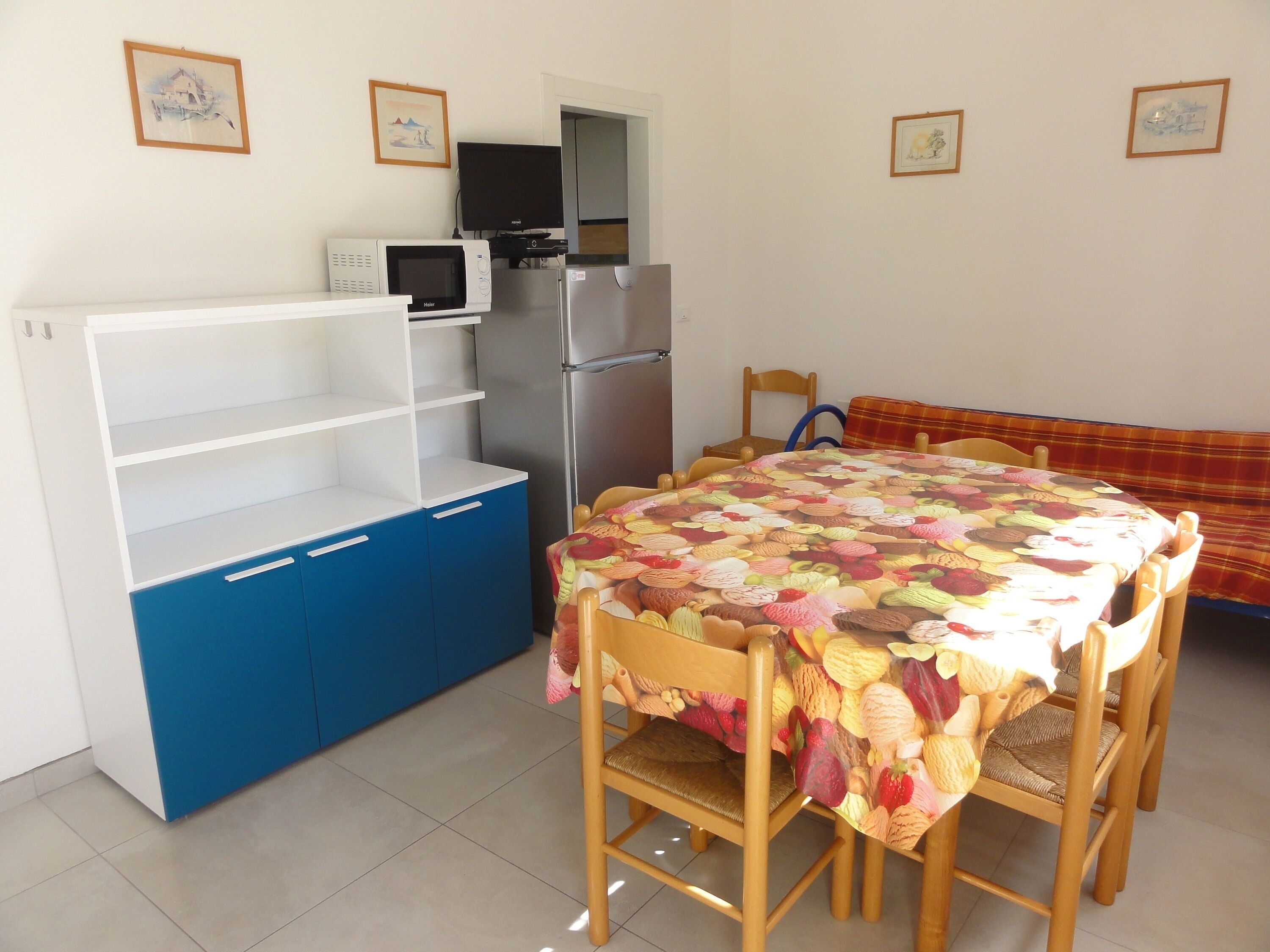Eine kleine Küche mit Mikrowelle, Herd und Kühlschrank. Ein Esstisch mit Stühlen und einer bunten Tischdecke steht bereit.