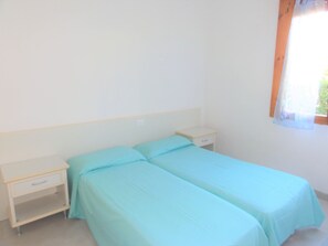 Una habitación pequeña y sencilla con dos camas individuales, una mesita de noche y una ventana con cortinas.