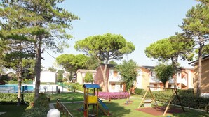 A playground with a slide and swings, surrounded by well-maintained grass and trees, with buildings in the background. - Bright Flat With Pool and Balcony (San Michele al Tagliamento)