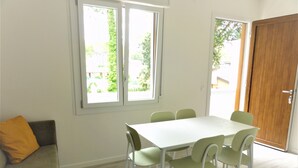 A dining area with a white table and green chairs, a wooden door, and a window with a view of greenery. - Bright Flat With Pool and Balcony (San Michele al Tagliamento)