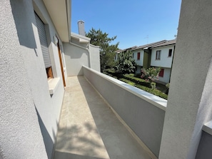 Un balcon étroit doté d’un sol carrelé, de murs blancs et offrant une vue sur les maisons voisines et la verdure.