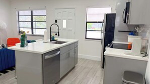 A compact kitchen with a dishwasher, a microwave, and a coffee machine. There are two windows with blinds, a door, and a small seating area with a chair and a cushion. - After Hours - Wilton Drive and Jacuzzi (Wilton Manors)