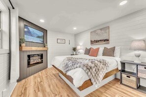 Ein modernes Schlafzimmer mit einem großen Bett, einem an der Wand befestigten Fernseher, einem Kamin und einem Nachttisch mit einer Lampe.