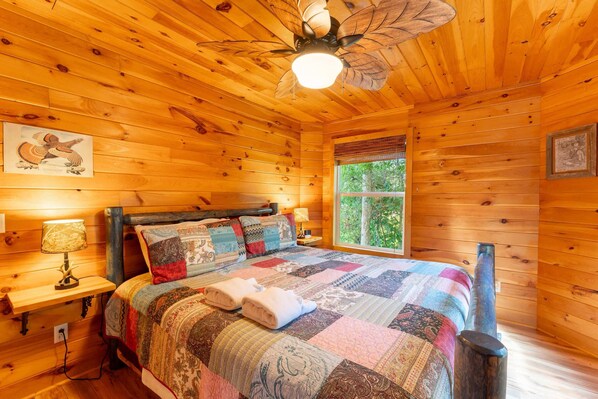 A wooden cabin bedroom with a bed, a nightstand, a lamp, a window, and a ceiling fan.