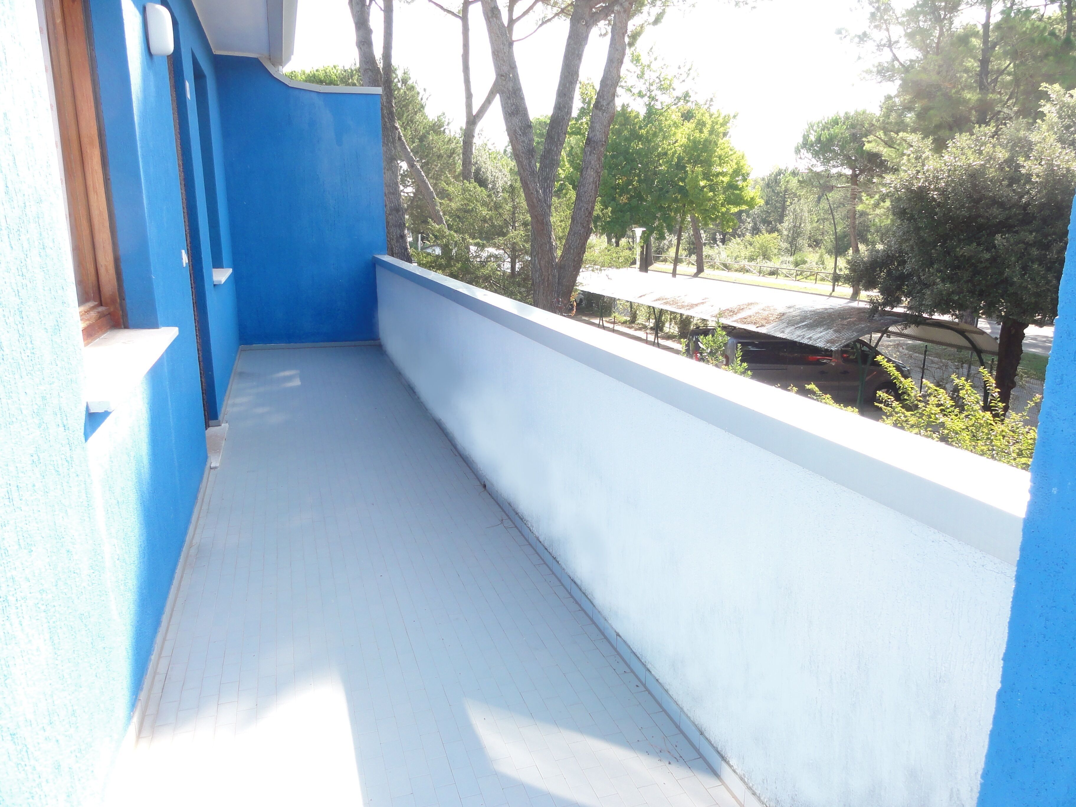 Ein Balkon mit blauer Wand und weißem Boden, der auf einen Parkplatz mit Bäumen und einer Überdachung blickt.