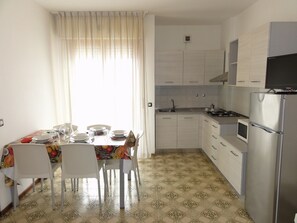 A compact kitchen with white cabinets, a stainless steel refrigerator, and a dining area with a table set for four. - Bright Flat Close to the Beach (San Michele al Tagliamento)