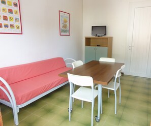 A room with a wooden table, white chairs, a red sofa, a television on a cabinet, and a door. - Comfy Apartment With Pool-beahost (San Michele al Tagliamento)