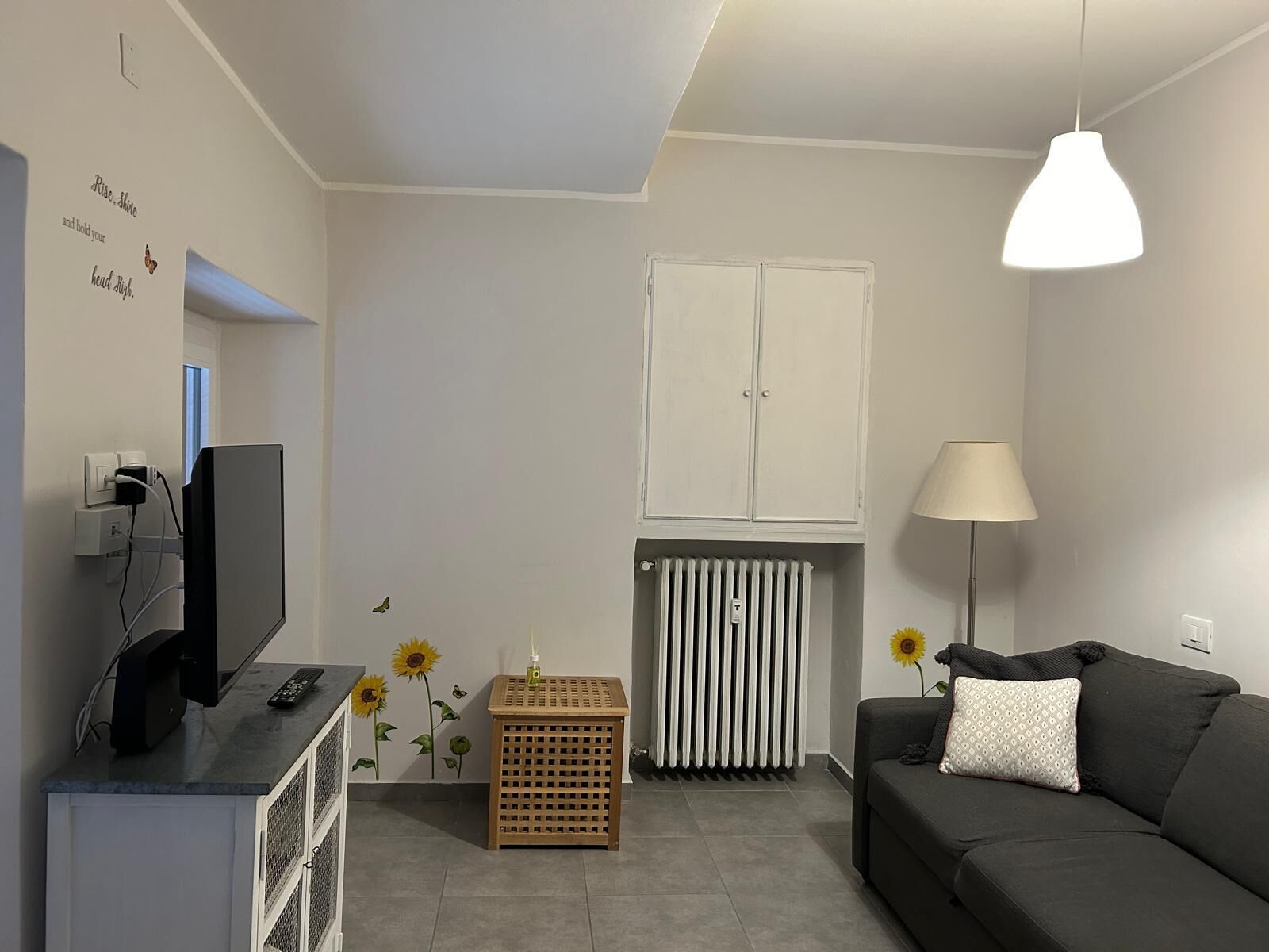 A modern living room with a grey sofa, a TV, a wooden side table, a radiator, and a white cabinet.
