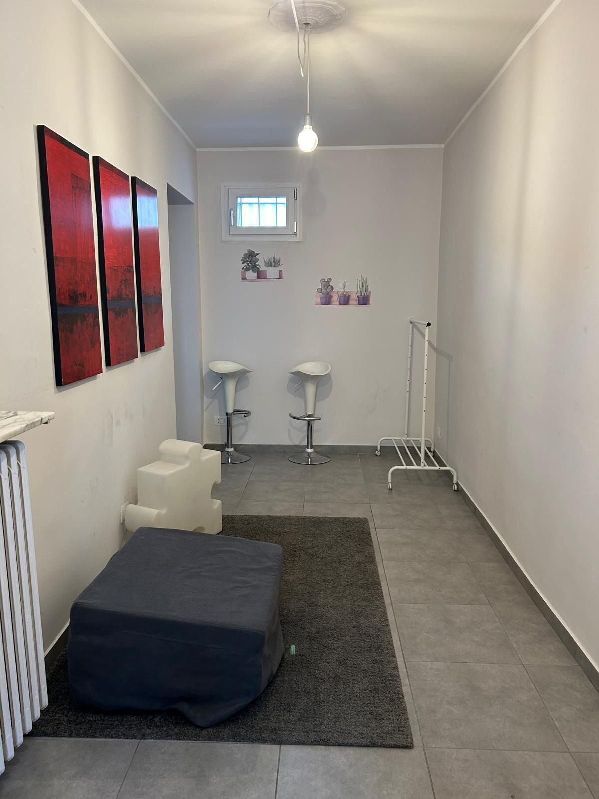 A small, modern room with a bed, two white bar stools, a radiator, and a ceiling light.