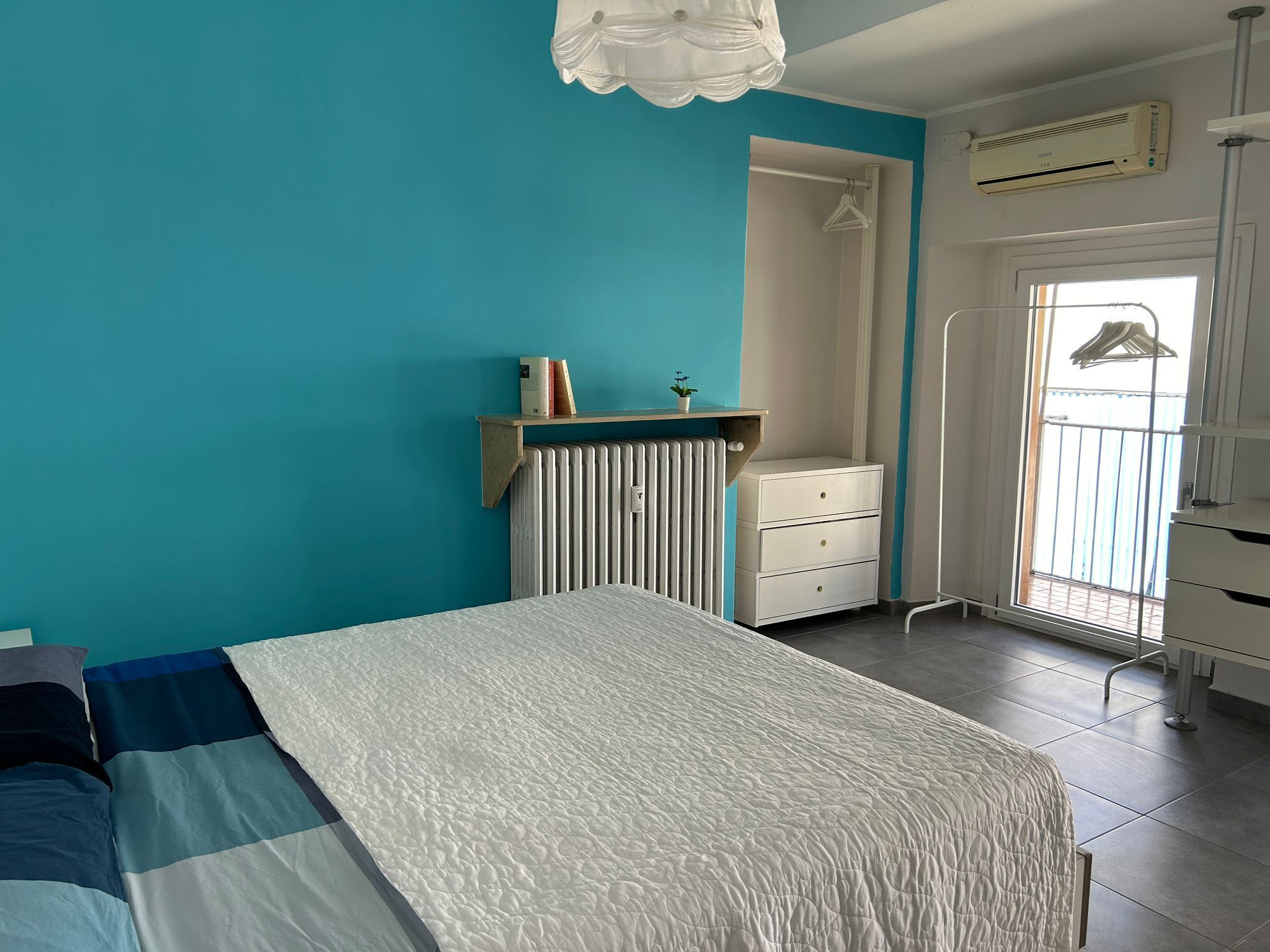 A bedroom with a bed, a radiator, a white dresser, a hanging clothes rack, and a window with a view of a balcony.