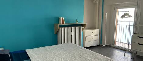A bedroom with a bed, a radiator, a white dresser, a hanging clothes rack, and a window with a view of a balcony.