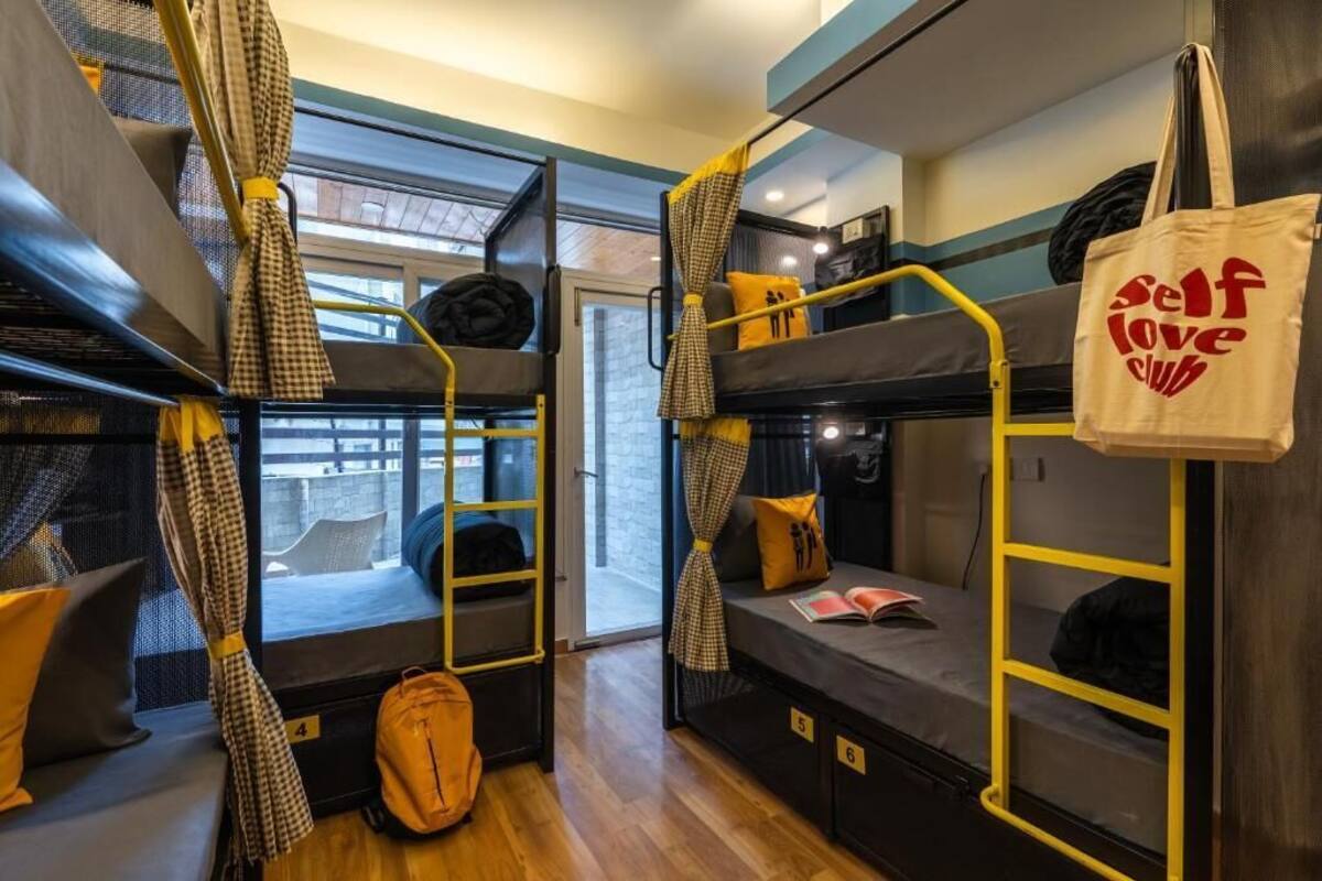 A dormitory room with bunk beds, a window with curtains, a backpack on the floor, and a tote bag with "self love club" text.