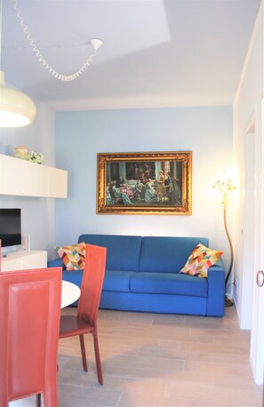 Un soggiorno con un divano blu, un quadro appeso al muro, un tavolo da pranzo con sedie e una lampada.