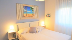 A neatly made bed with a pillow, a bedside lamp, a nightstand, and a painting of a beach scene on the wall. - Ground-floor Flat With Pool (San Michele al Tagliamento)
