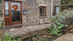 Una casa in pietra con una porta e una finestra in legno, un piccolo portico e un giardino con piante e un recinto di legno.