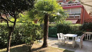 A patio area with a table and chairs, surrounded by greenery and a building in the background. - Apartment With Pool Access Near Beach (San Michele al Tagliamento)