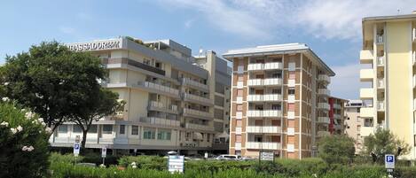 A multi-story residential building with balconies, a parking sign, and a tree in the foreground.