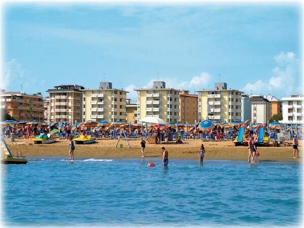 A crowded beach with people swimming and sunbathing, and buildings in the background.