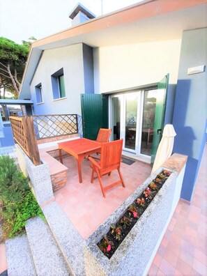 A patio with a table and chairs, a small garden bed with flowers, and a sliding glass door leading to a covered area. - Comfy Villa With Garden (San Michele al Tagliamento)