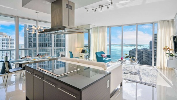 Cozinha moderna com ilha de vidro, área de jantar com sofá e mesa, e grandes janelas com vista para a cidade.