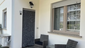 A small outdoor seating area with two wicker chairs and a table, a window with blinds, and an air conditioning unit on the wall. - Bibione Apartment Close to Promenade (San Michele al Tagliamento)
