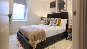 A bedroom with a bed, a bedside table, a window, and a wall-mounted picture. - Streatham Hill Modern Flat with Balcony (London)