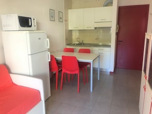 Eine kompakte Küche mit weißen Schränken, einer Mikrowelle und einem Kühlschrank. Ein Esstisch mit roten Stühlen steht daneben.