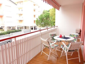 Un balcón con sillas y una mesa blancos, con vista a una calle con coches aparcados y edificios.