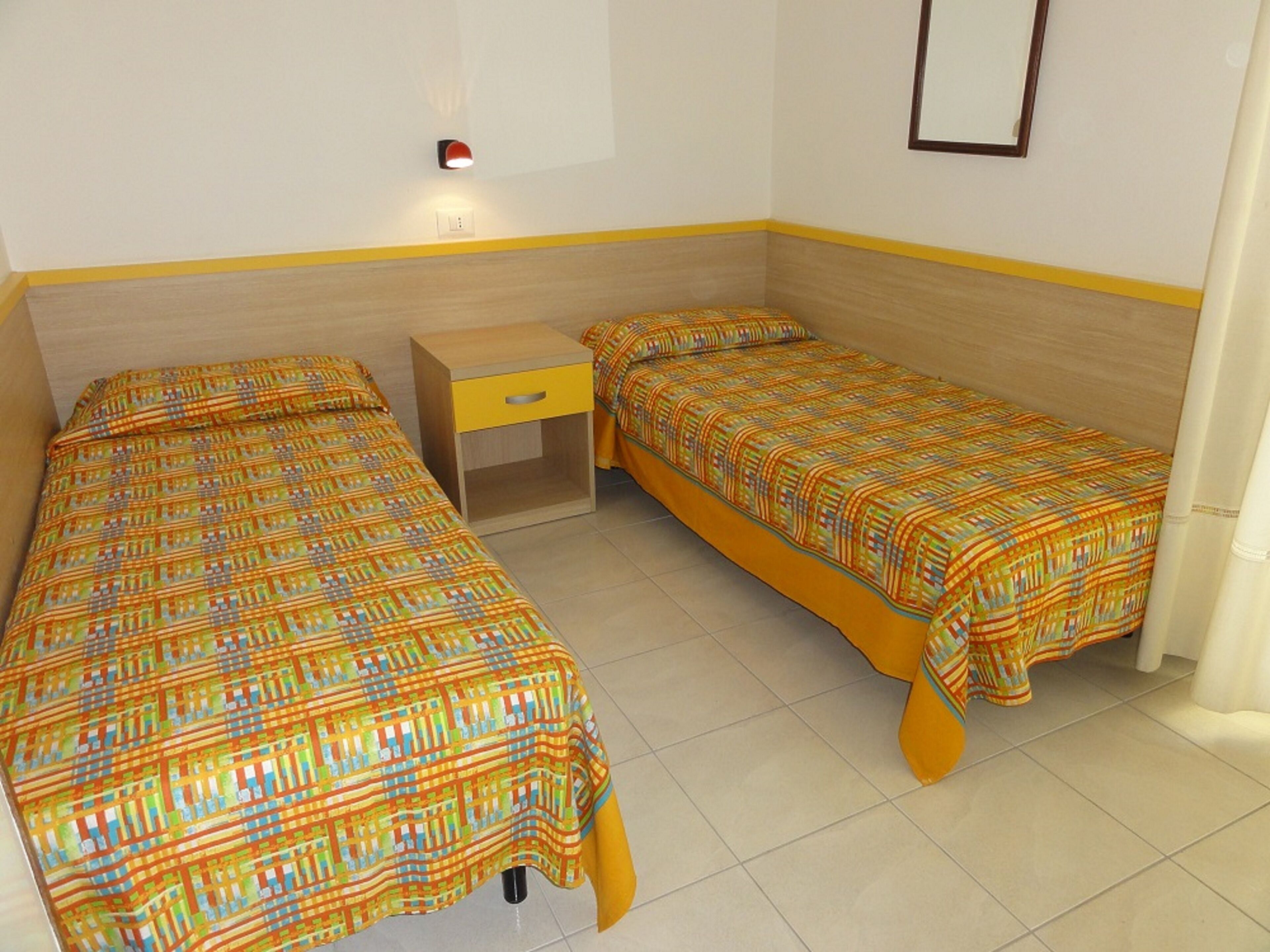 A room with two beds, a small bedside table, a wall-mounted light, and a framed picture.