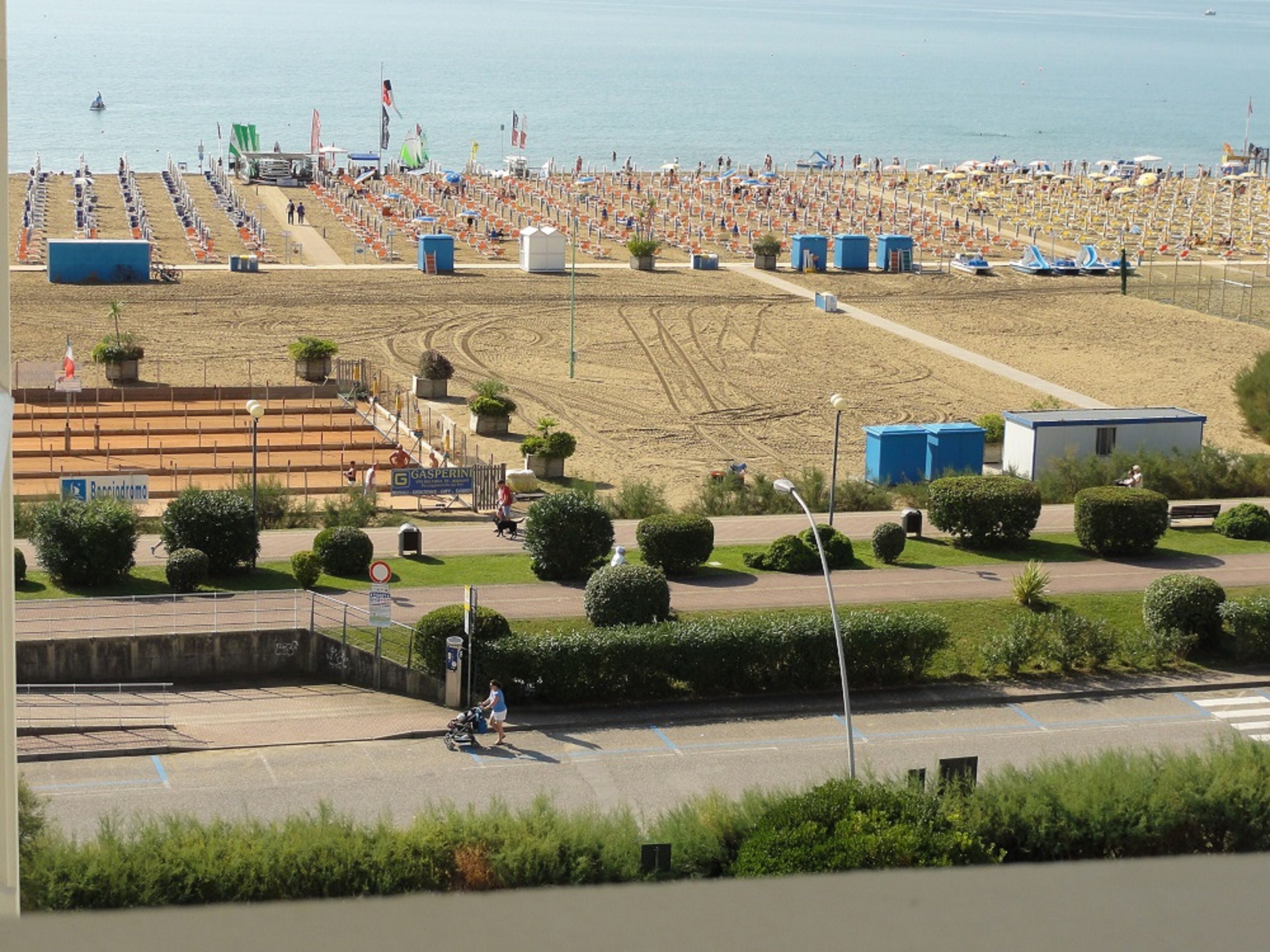 Een strand met rijen ligbedden en parasols, een parkeerplaats en een weg met een zebrapad.