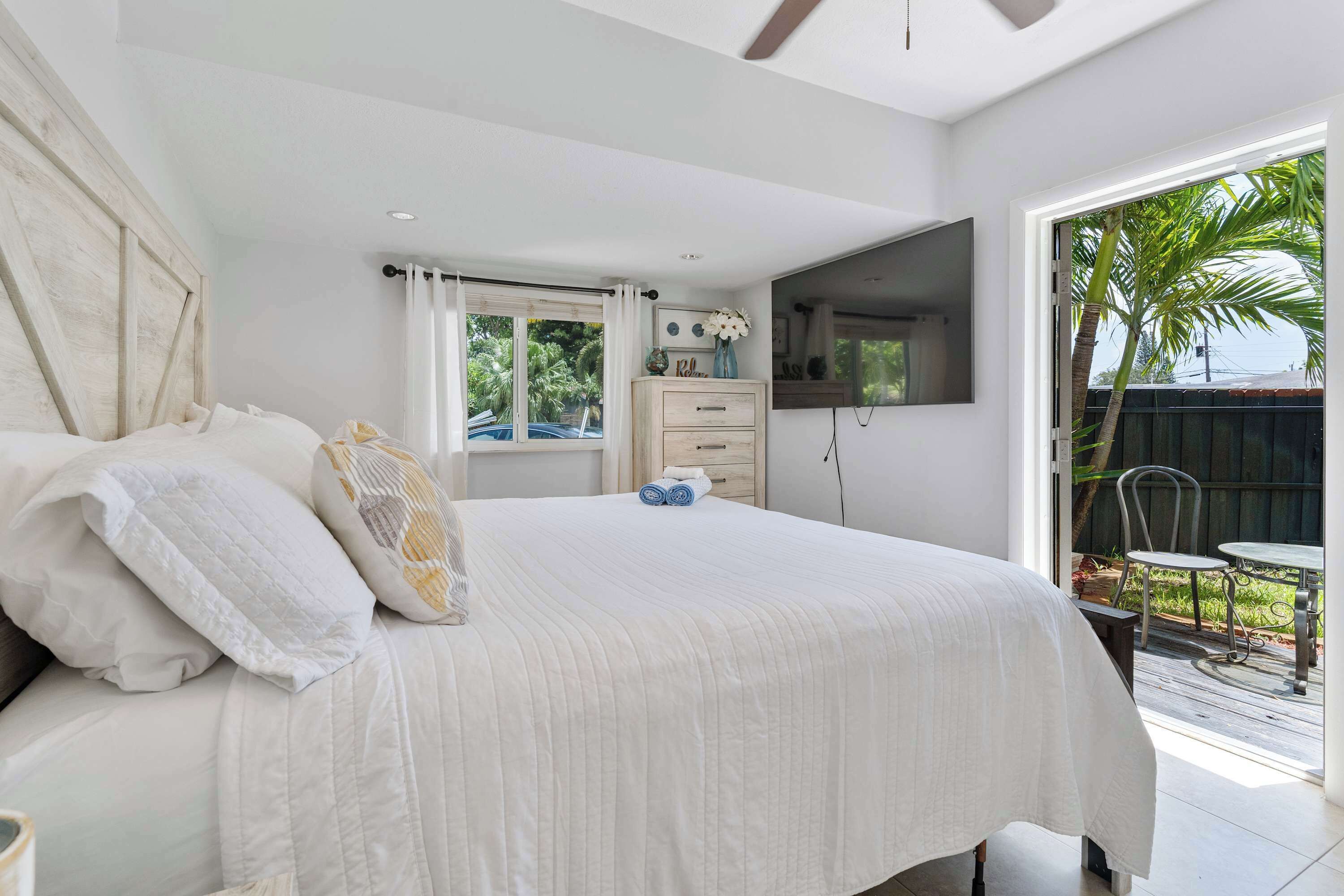 A bedroom with a large bed, a TV mounted on the wall, a window with curtains, and a view of the outdoors.