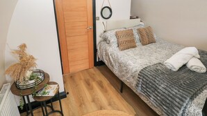 A bedroom with a wooden door, a bed with patterned bedding, a bedside table with a plant, and a small round table with a book. - Cwtch Bach (Aberdare)