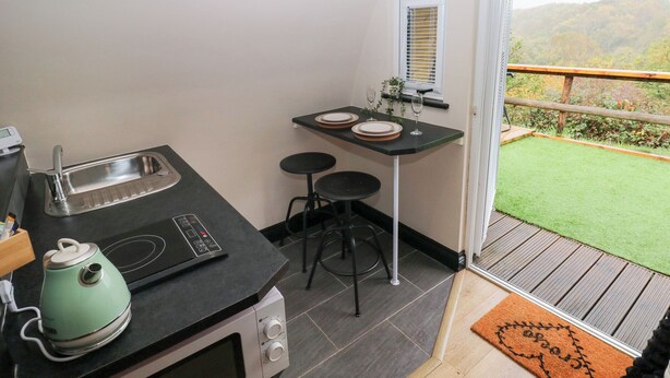 A compact kitchen with a sink, a kettle, and a microwave. There is a small table with two stools and a window with blinds. The room has a view of a green lawn and hills. - Cwtch Bach (Aberdare)
