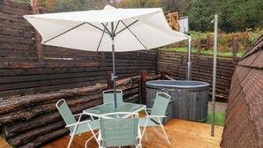 An outdoor patio with a wooden deck, a table with chairs, a white umbrella, and a hot tub. - Cwtch Bach (Aberdare)