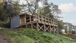 A wooden deck structure with a metal building adjacent to it, set on a grassy hill with trees in the background.