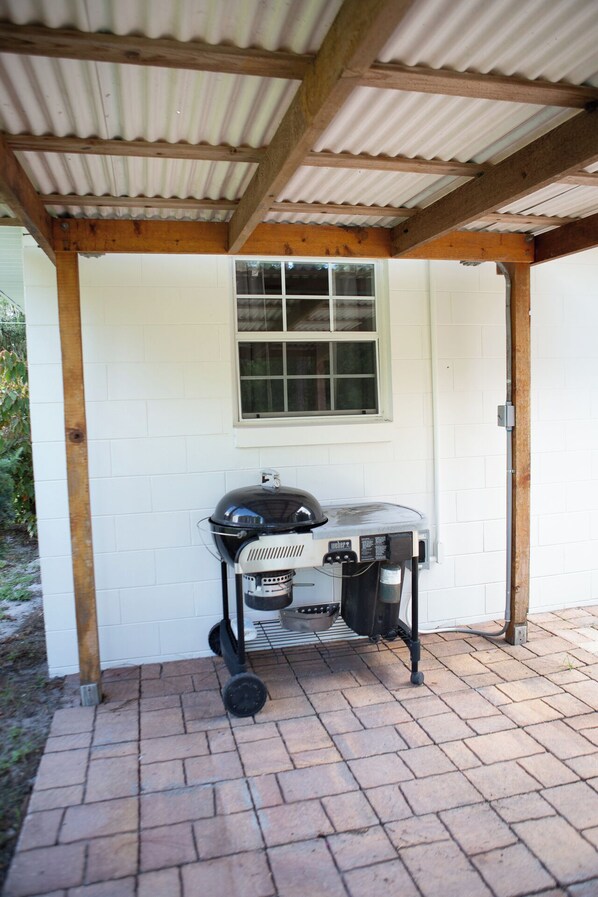 Un espace extérieur abrité, doté d’un barbecue Weber sous un toit en tôle ondulée, de murs en briques blanches et d’une fenêtre.