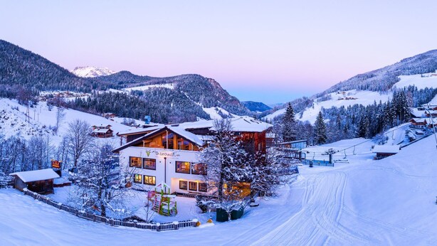 A snow-covered mountain resort with a prominent lodge featuring multiple windows and a ski lift visible in the background. - Familienhotel Unterreith (Forstau)