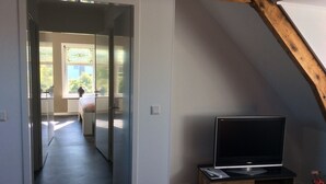 Et værelse med et fjernsyn på et træstel, en dør der fører til et andet rum, og et vindue med udsigt over grøn vegetation udendørs.