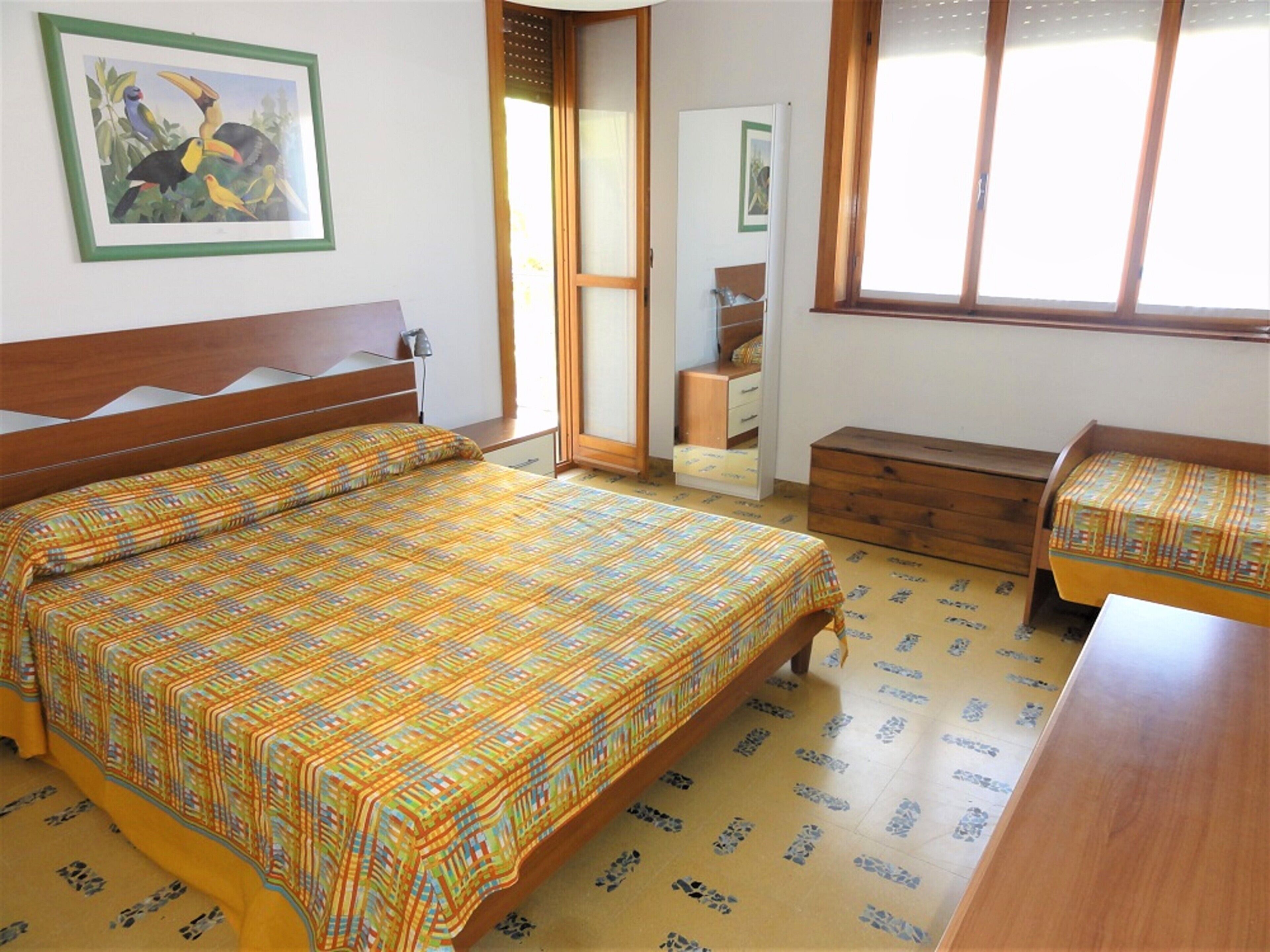 Ein Schlafzimmer mit einem hölzernen Bett, einer kleinen Holzbank, einem Schminktisch und einem Fenster mit Blick ins Freie.
