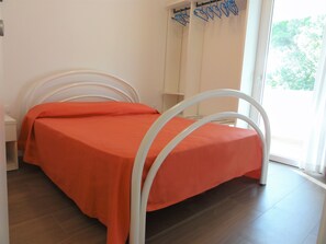 Un lit simple avec un cadre en métal blanc et une couvre-lit orange, dans une pièce dotée d’un plancher en bois et d’une fenêtre avec des rideaux.