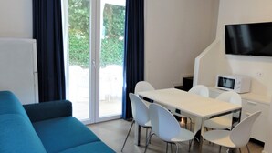 A modern living room with a blue sofa, a dining table with white chairs, a flat-screen TV, and a microwave. - Modern Terraced House (San Michele al Tagliamento)